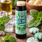 Lip balm labeled 'Mint Kiss Me' surrounded by mint leaves, a bottle of oil, and candy on a wooden surface.